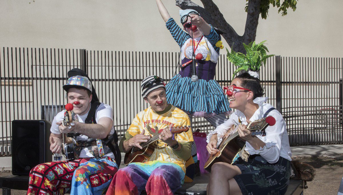 Four clown musicians play guitars and ukuleles outdoors while a fifth performs standing behind them.