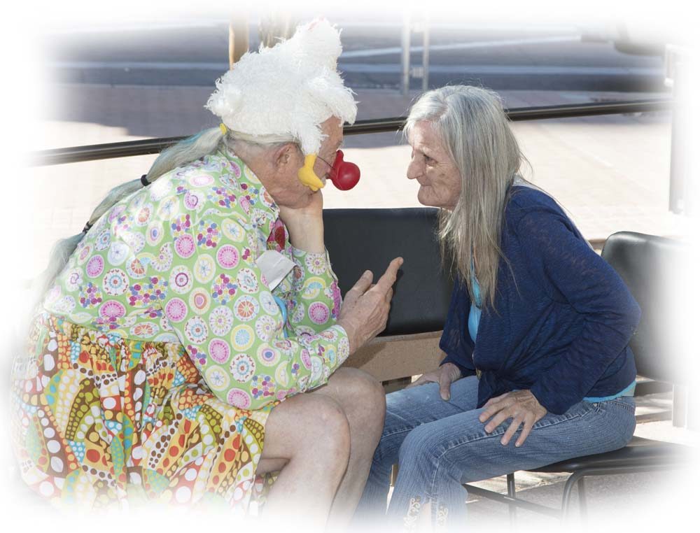 A red-nose clown leans in to listen to a woman seated on a public bench.